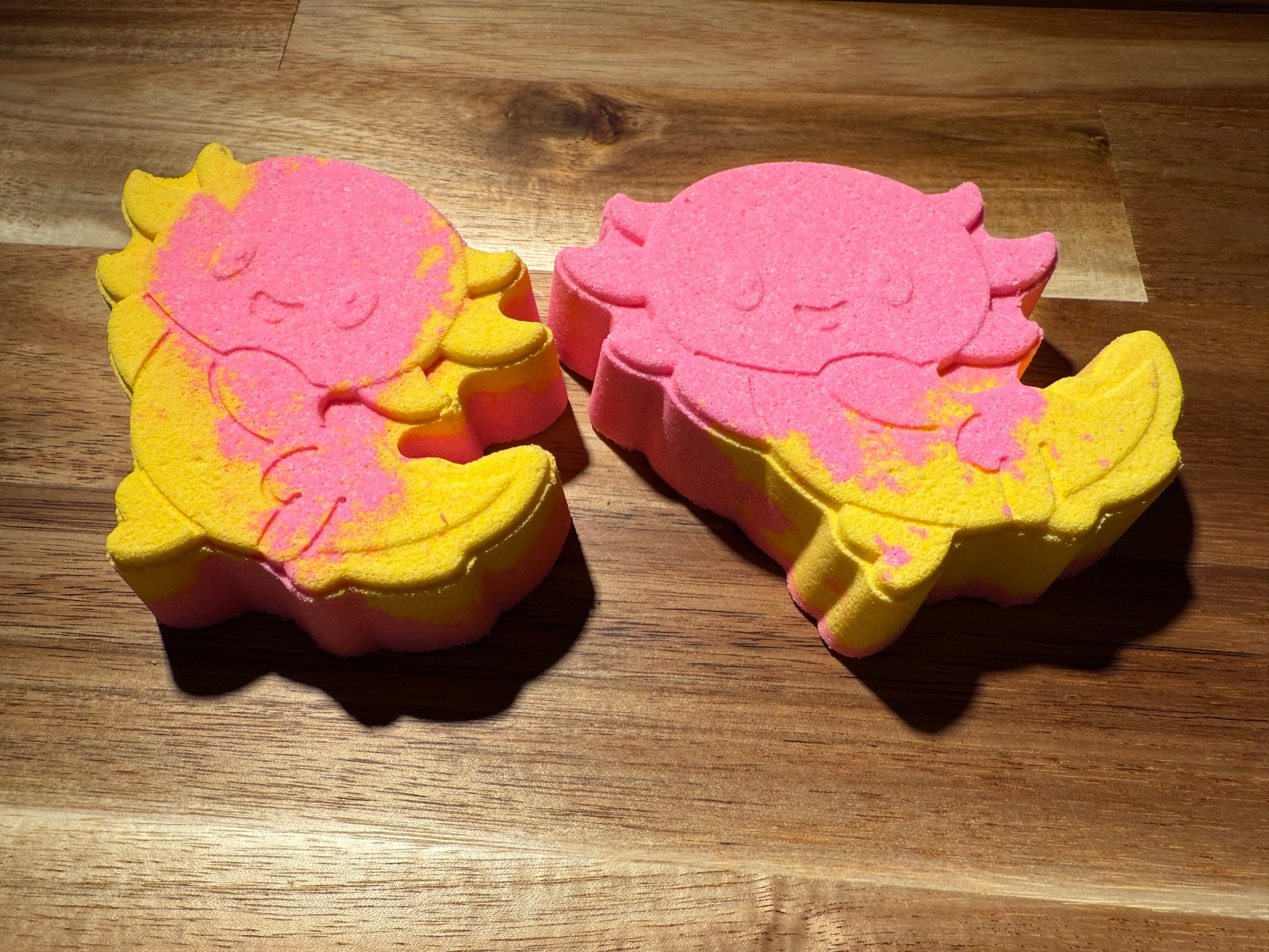 Two pink and yellow bath bombs shaped like a seashell and a crab on a wooden surface.